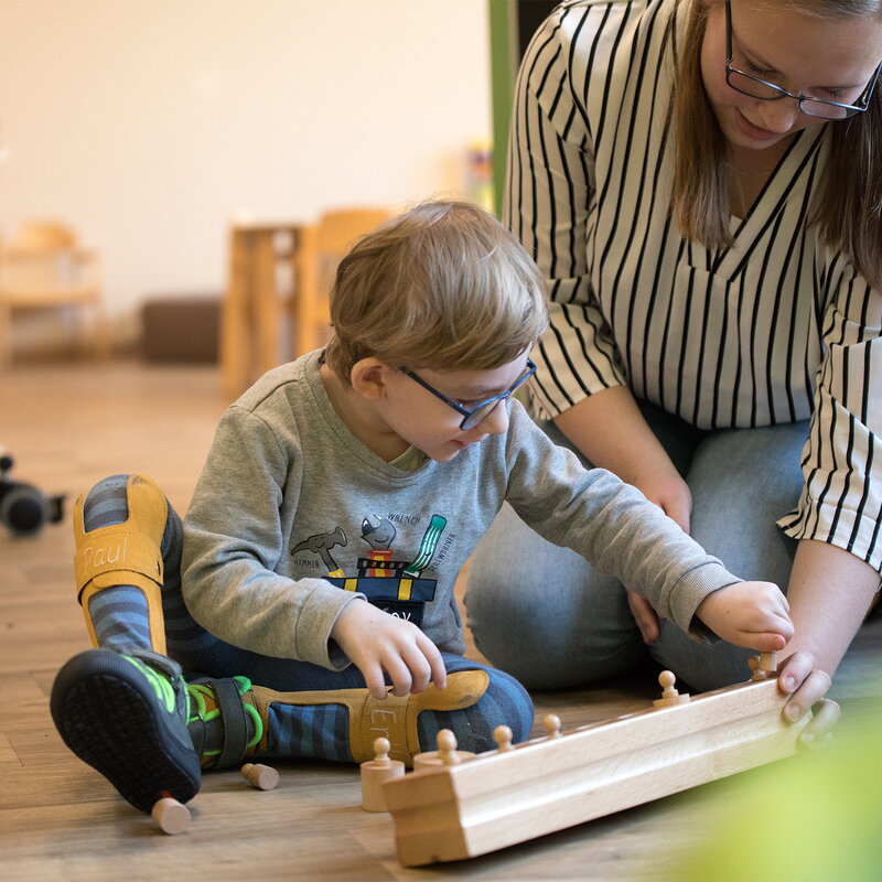 Ein kleiner Junge mit Beinschienen sitzt auf dem Boden und steckt Holzzylinder in ein Montessori-Spielzeug. Eine Erzieherin kniet neben ihm und unterstützt ihn aufmerksam.