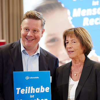 Ein Mann und eine Frau halten ein blaues Schild vor unscharfem Hintergrund