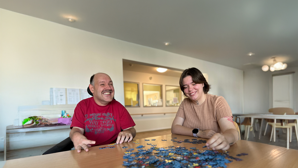 Ein Mensch mit Unterstützungsbedarf und eine junge Frau sitzen gemeinsam am Tisch und puzzeln.