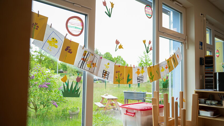Frühlingshaft geschmücktes Kita-Fenster mit Blick ins Grüne. Quer über die Scheibe spannt sich eine Leine mit bunten Kinderbildern, davor stehen umgedrehte Holzstühle.