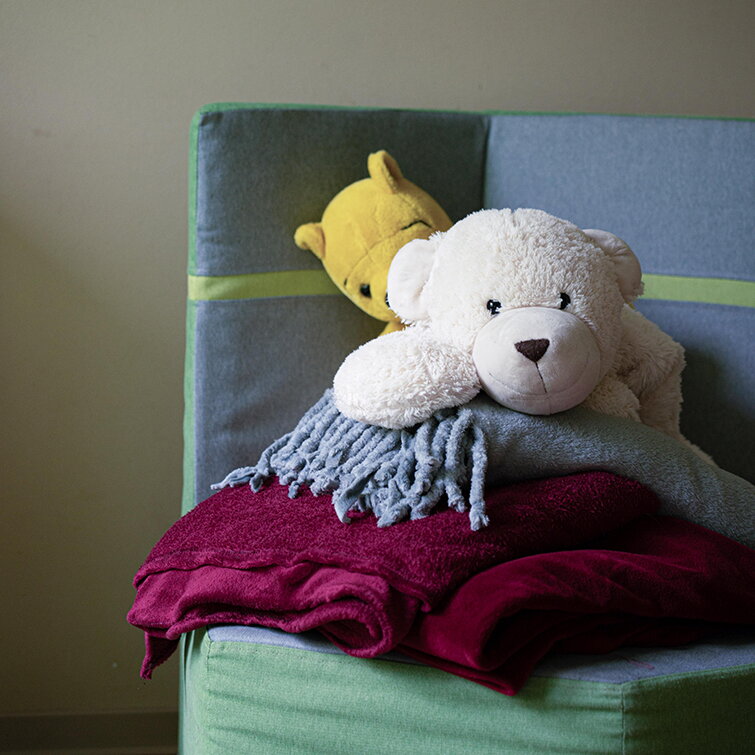 Ein weißer und ein gelber Kuschelbär liegen gemütlich auf einem Stapel weicher, bunter Decken. Sie kuscheln sich in die Ecke eines grün-grauen Polstersessels in der Kita.