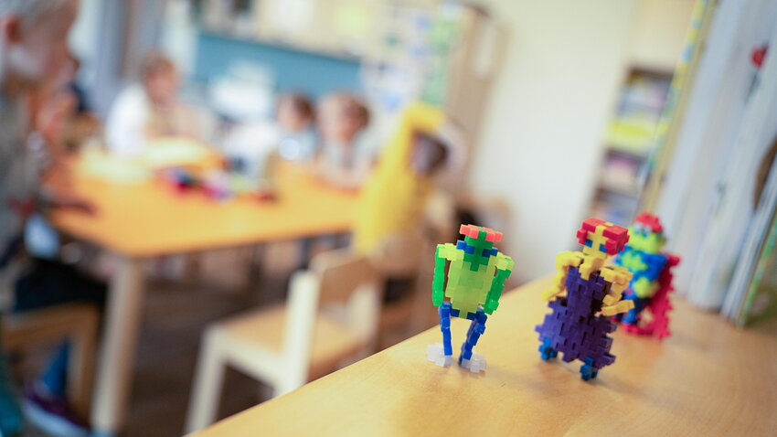 Kleine, bunte Spielfiguren aus Steckbausteinen stehen fokussiert im Vordergrund. Der Hintergrund zeigt verschwommen Kinder an einem Tisch in einem hellen Gruppenraum.