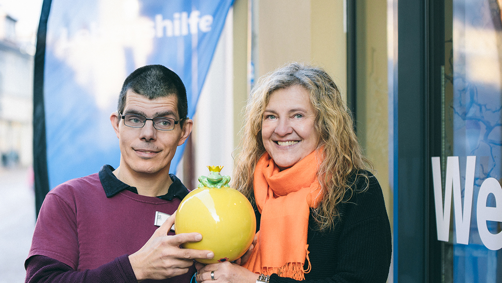 Zwei Mitarbeiter aus dem Inklusiven Team des Werkstattladens. Sie halten gemeinsam eine gelbe Keramik mit einem Froschkönig darauf.