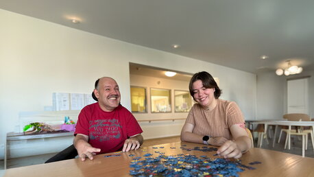 Ein Mensch mit Unterstützungsbedarf und eine junge Frau sitzen gemeinsam am Tisch und puzzeln.