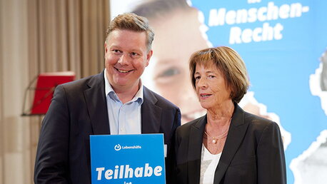 Ein Mann und eine Frau halten ein blaues Schild vor unscharfem Hintergrund
