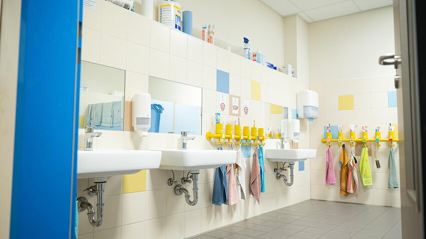 Ein gefliester Kita-Waschraum mit mehreren niedrigen Waschbecken und Spiegeln. An gelben Hakenleisten hängen bunte Zahnputzbecher und Handtücher für die Kinder bereit.