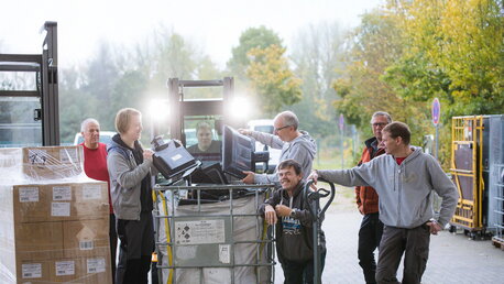 Eine Gruppe von Männern steht vor einem Gabelstapler und sortiert Elektroschrott in eine große Box.