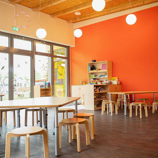 Ein heller Kita-Gruppenraum mit einer leuchtend orangefarbenen Wand, Holzmöbeln und runden Hängelampen. Große Glastüren führen nach draußen auf das Außengelände.