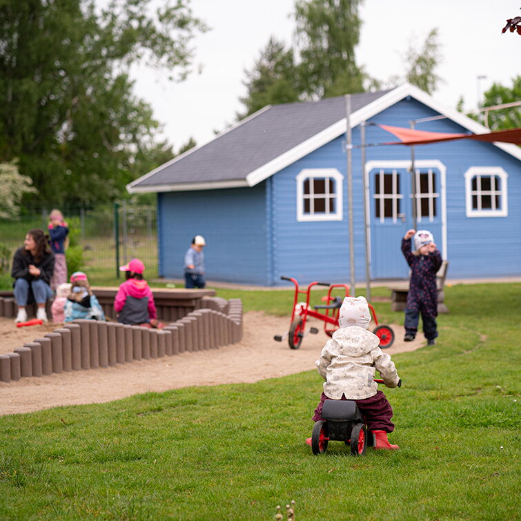 Ein Kleinkind fährt im Vordergrund auf einem Spielauto über die Wiese einer Kita. Dahinter spielen weitere Kinder im Sandkasten und im Garten vor einem blauen Holzhaus.