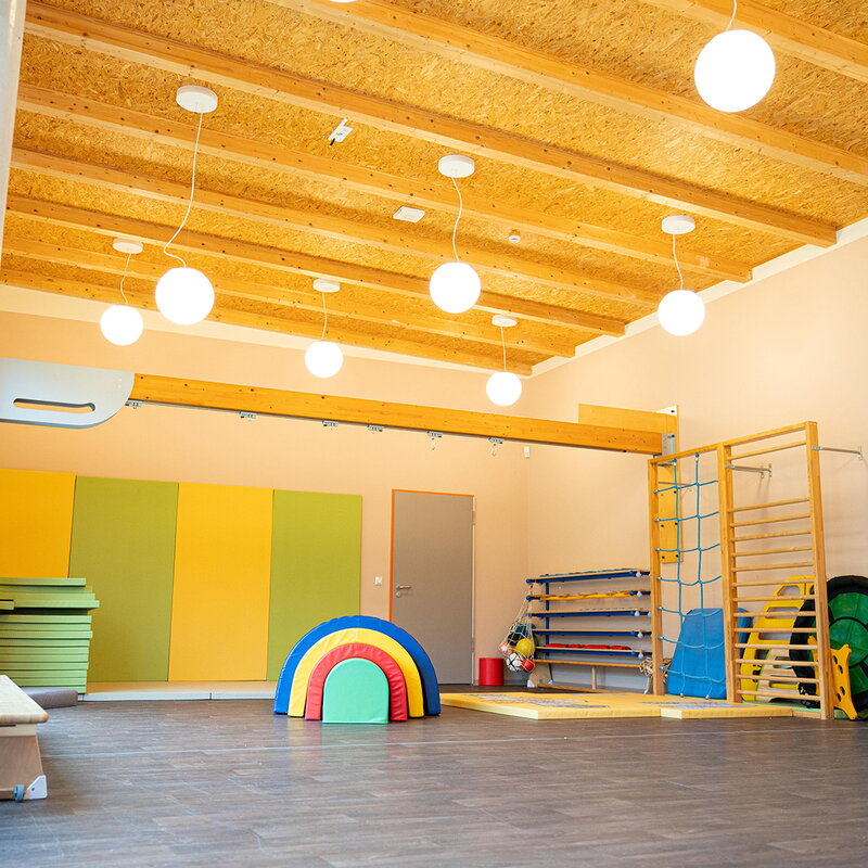 Ein großer, heller Turnraum der Kita mit hoher Holzdecke und runden Pendelleuchten. Im Raum verteilt stehen Klettergerüste, bunte Weichbodenmatten und Schaumstoff-Spielelemente.