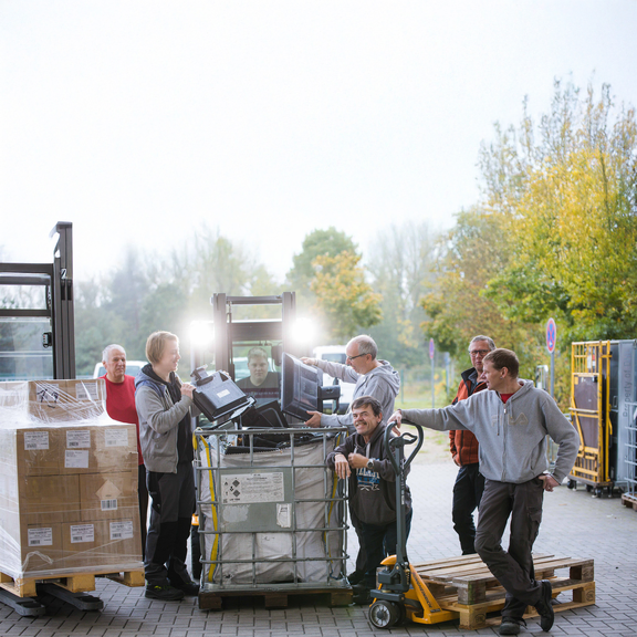Eine Gruppe von Männern steht vor einem Gabelstapler und sortiert Elektroschrott in eine große Box.