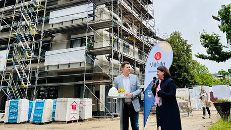 Ein Mann und eine Frau stehen beim Richtfest an einem Stehtisch vor einem mehrstöckigen Neubau