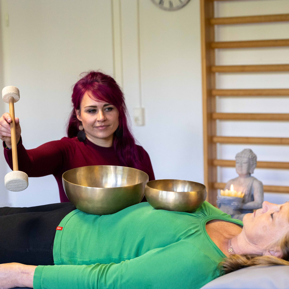 Eine junge Frau führt eine Klangschalentherapie an einer Patientin durch.
