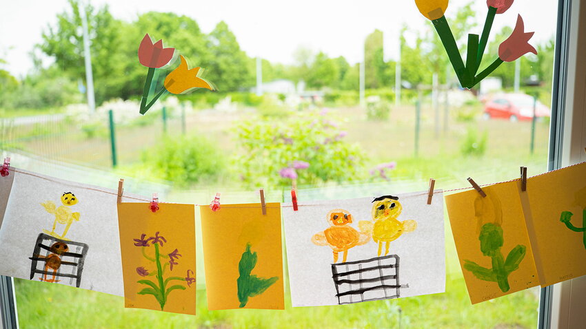 Bunte Kinderzeichnungen mit Küken- und Blumenmotiven hängen an einer Schnur vor einem Kita-Fenster. Auf der Scheibe kleben gebastelte Tulpen, dahinter ist eine Wiese sichtbar.