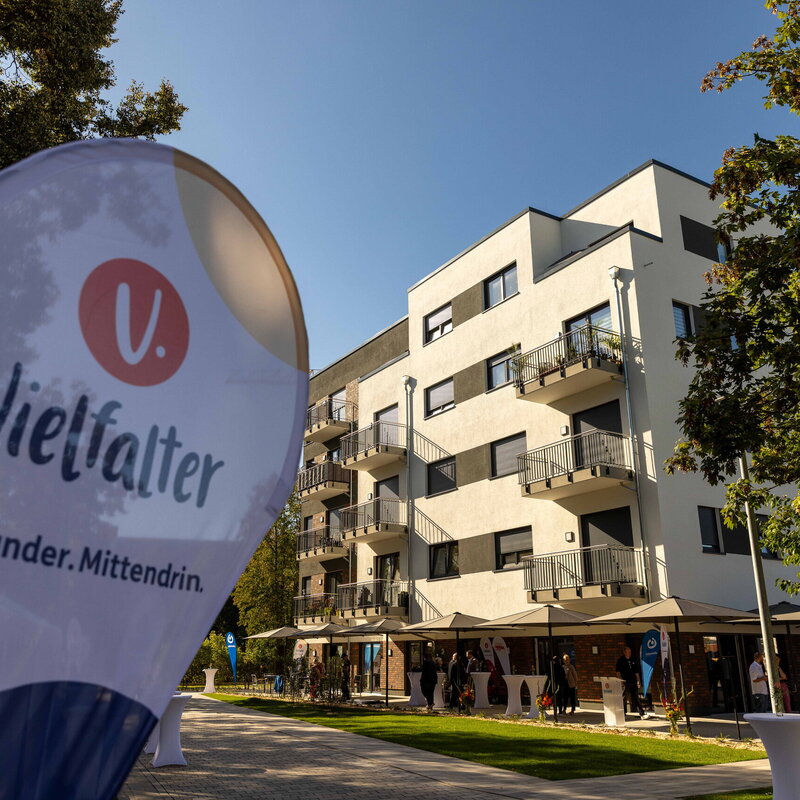 Eine Vielfalter Beachflag steht vor einem modernen Wohngebäude in der Abendsonne