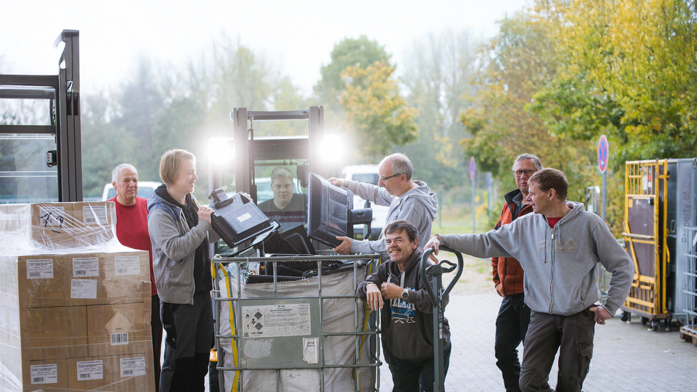 Eine Gruppe von Männern steht vor einem Gabelstapler und sortiert Elektroschrott in eine große Box.