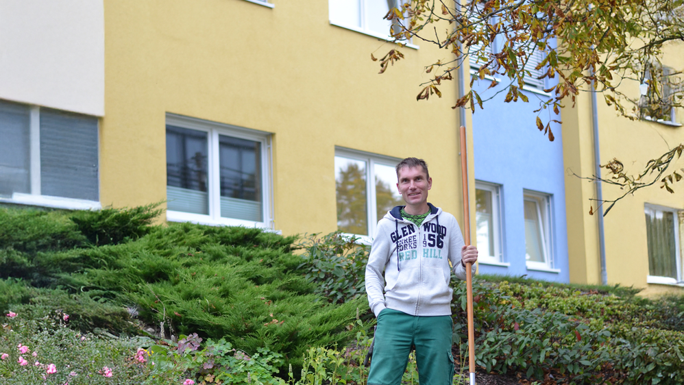 Mann in Arbeitskleidung steht mit einer Harke in der Grünanlage vor einem Neubaublock.