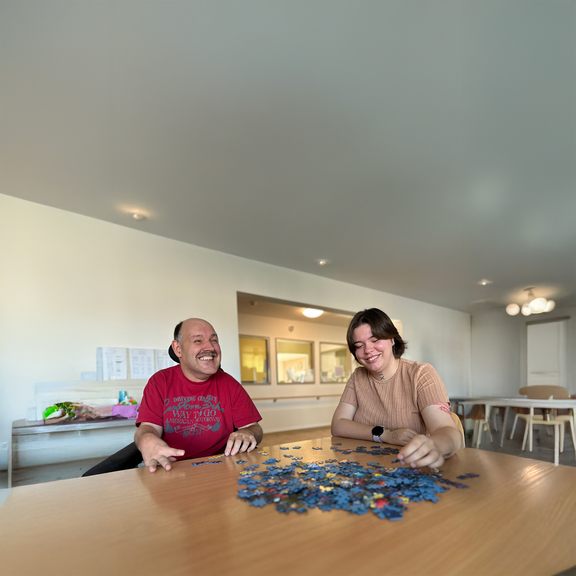 Ein Mensch mit Unterstützungsbedarf und eine junge Frau sitzen gemeinsam am Tisch und puzzeln.